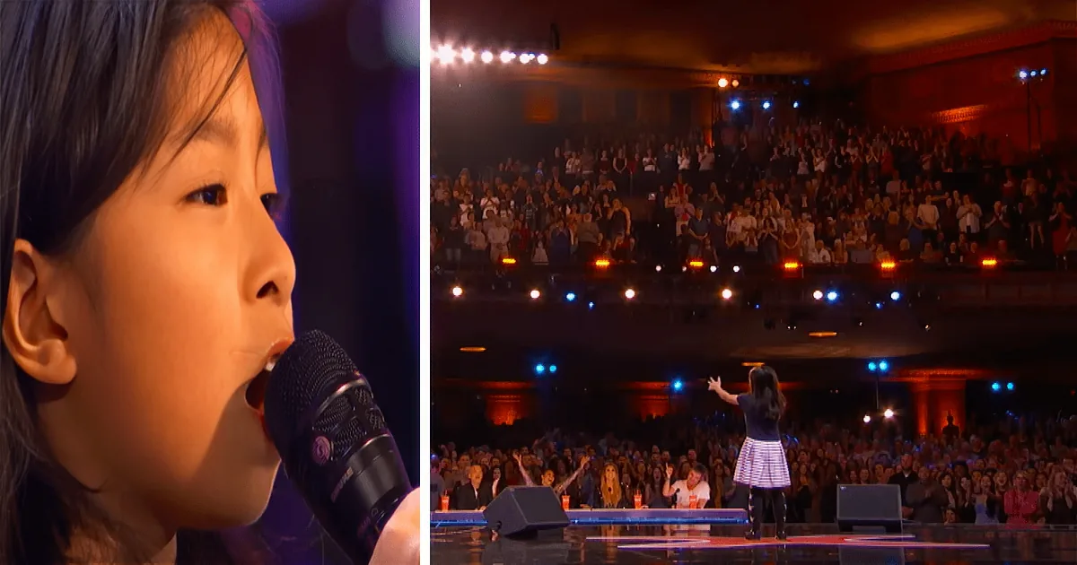 A young girl sings into a microphone on stage, with a large audience watching in a dimly lit theater.