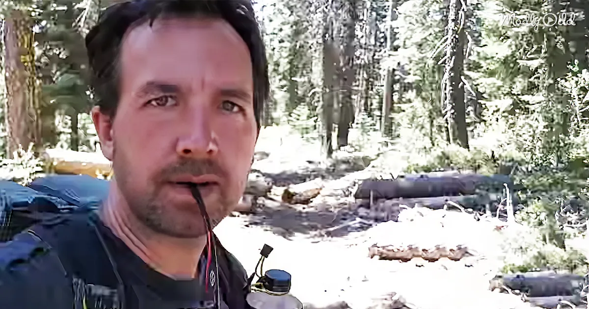 A man with a backpack and water bottle stands in a forested area with trees and rocks in the background.
