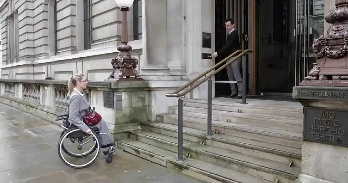 A woman in a wheelchair navigates a wet sidewalk near a building with steps and railings.