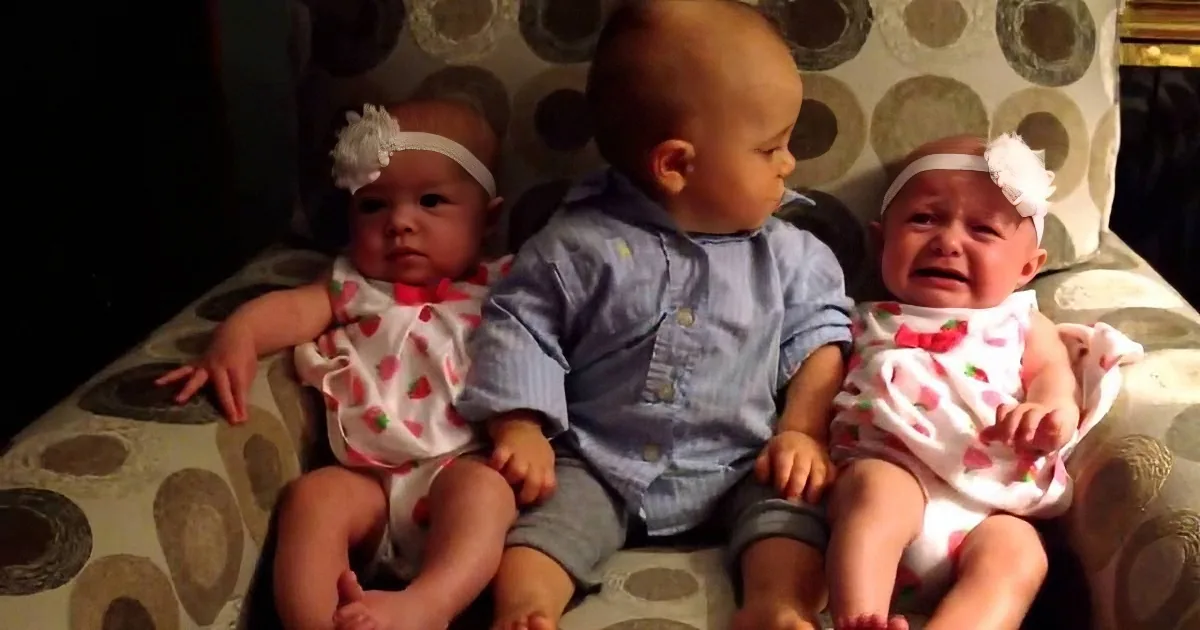 Three babies, two in matching outfits, sit together on a patterned couch, one crying.