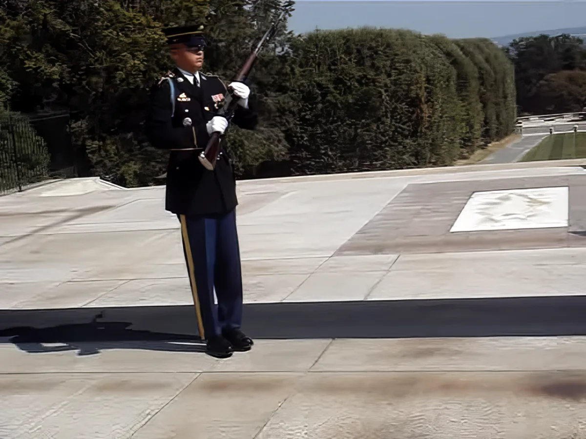 A soldier in formal uniform stands at attention, holding a rifle, with trees and a pathway in the background.