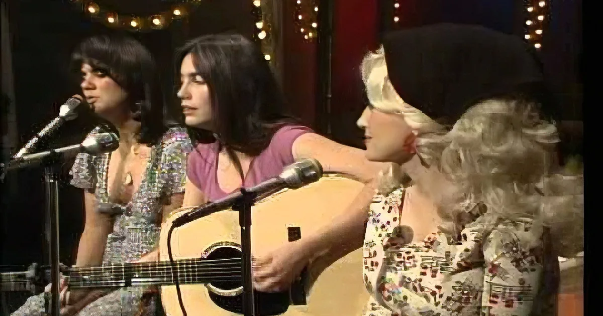 A woman in a floral dress plays guitar while another sings into a microphone.