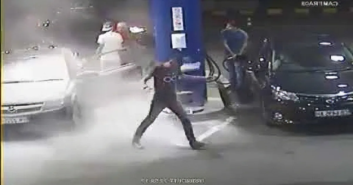A person in a black shirt runs past a gas pump, with a car and another individual in the background.