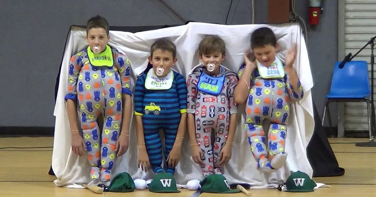 Four boys in pajamas and bibs stand on stage with pacifiers, smiling and waving.