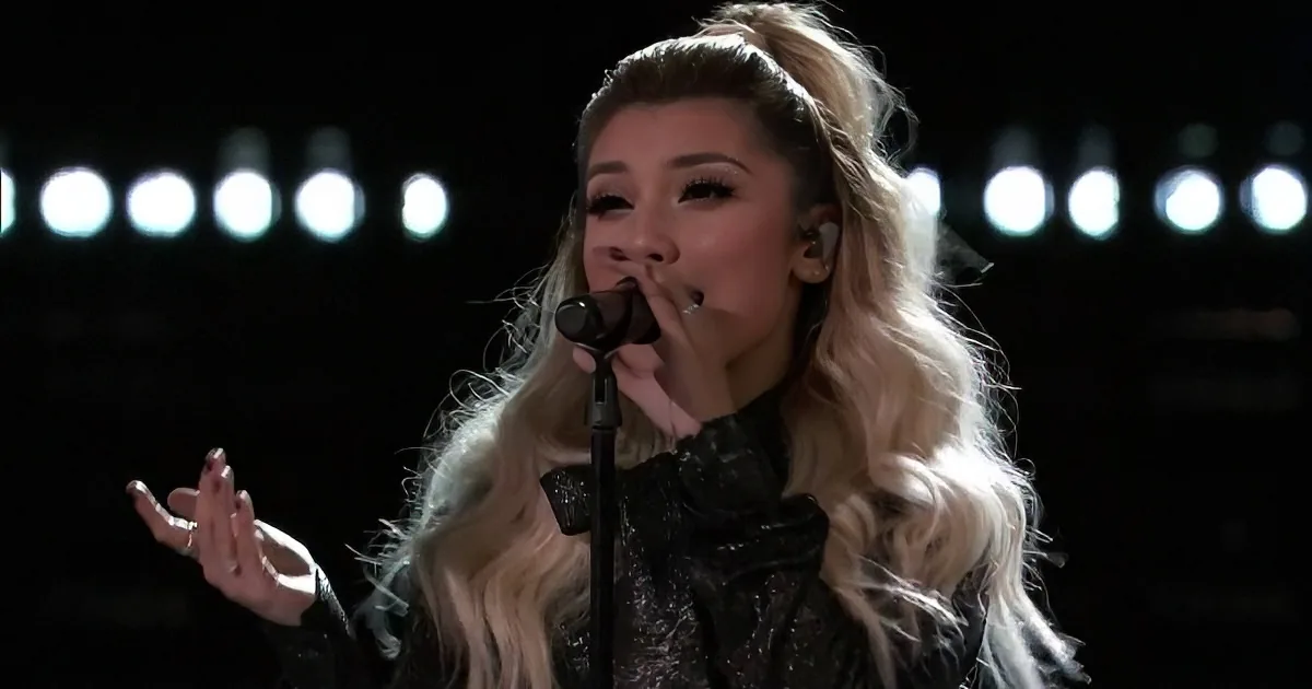 A blonde woman in a black outfit sings into a microphone, her hand raised, with bright lights in the background.