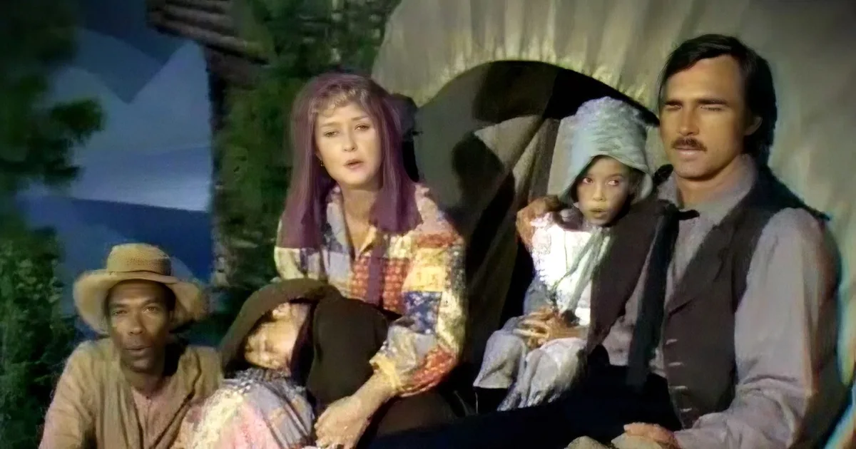 A woman in a patchwork dress and headscarf is seated, holding a child, with a man in a vest and hat beside her.