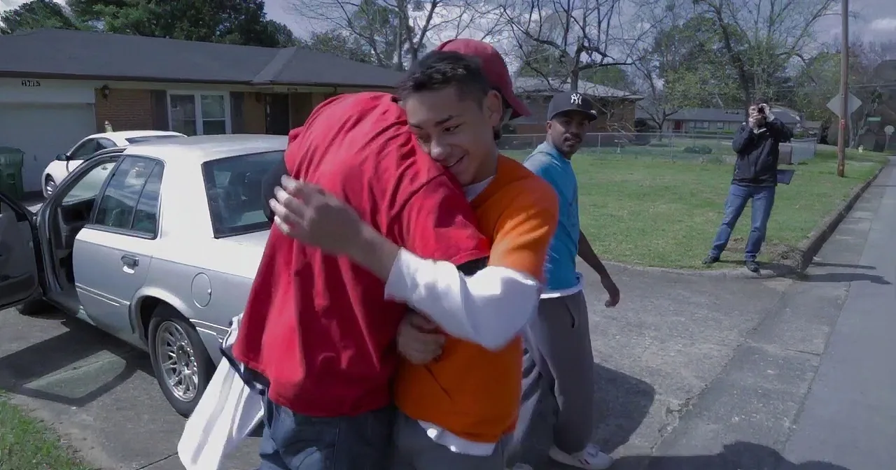 A boy in an orange shirt hugs another boy in a red hoodie on a sidewalk.
