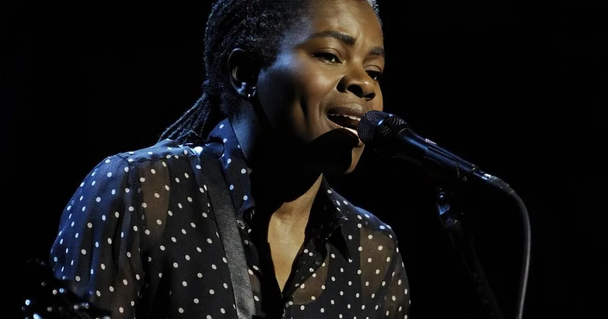 A person with dreadlocks sings into a microphone, wearing a polka-dot shirt against a dark background.