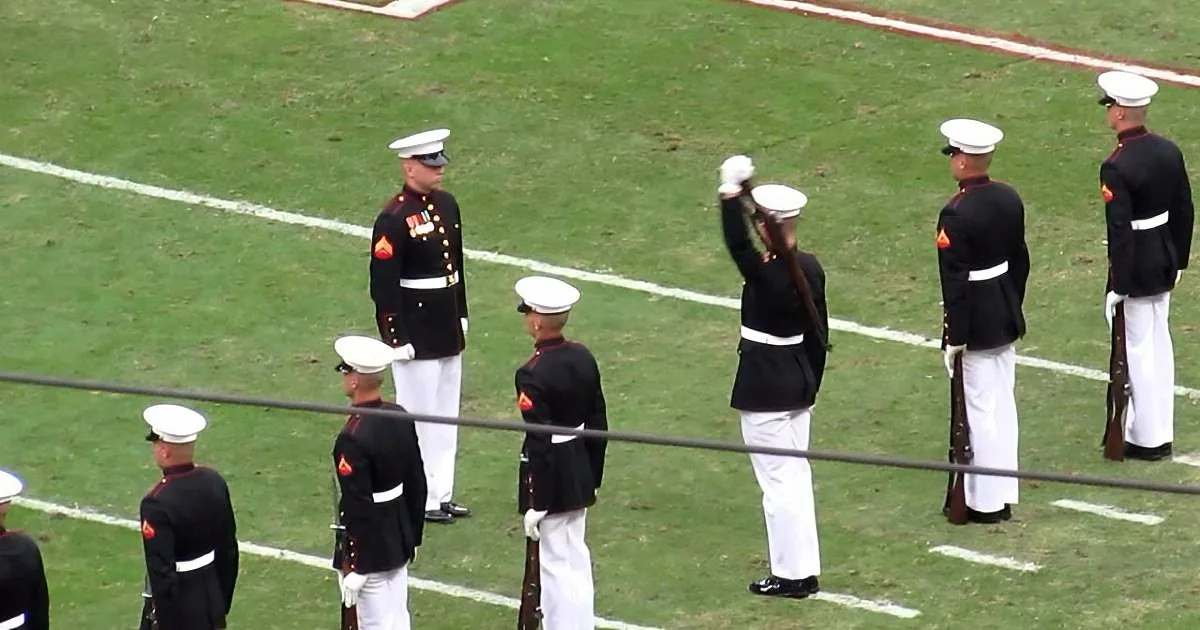 Marines in formal uniforms stand at attention on a field, saluting.