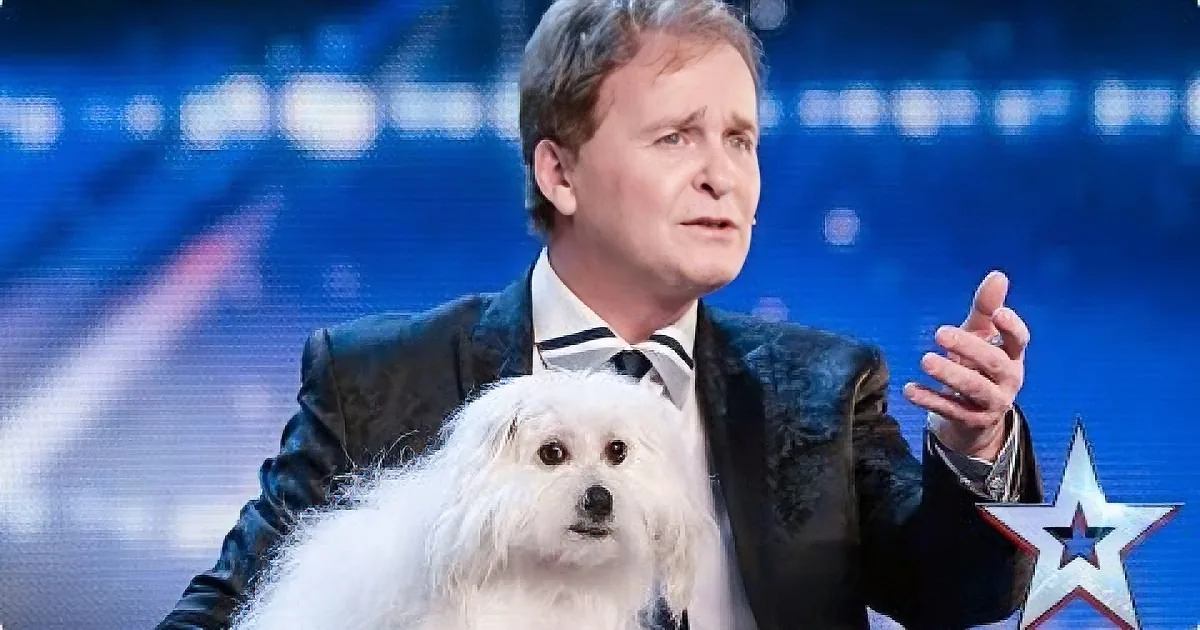 A man in a suit gestures while holding a fluffy white dog on stage.
