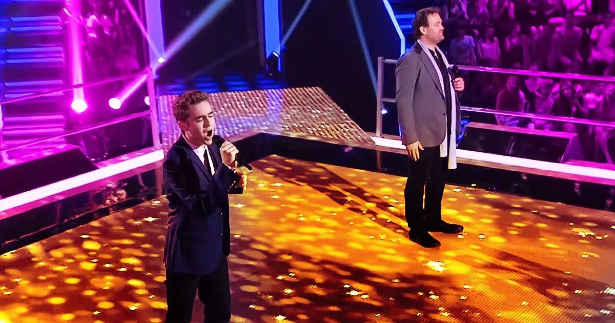 A man in a suit sings passionately on stage, illuminated by vibrant purple and gold lights.