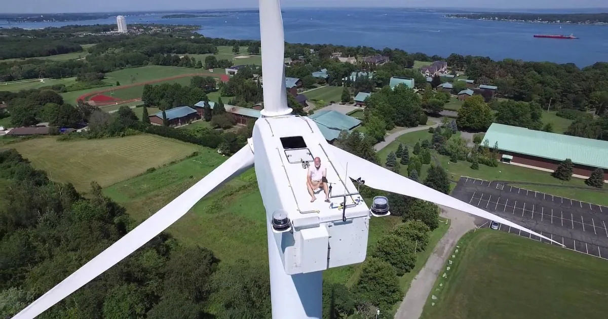 A person sits on the top of a wind turbine overlooking a lush green landscape and a body of water.
