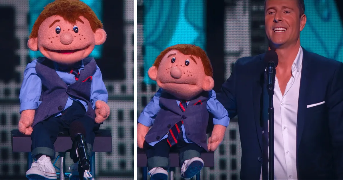 A puppet in a suit sits on a chair, next to a man in a suit holding a microphone.