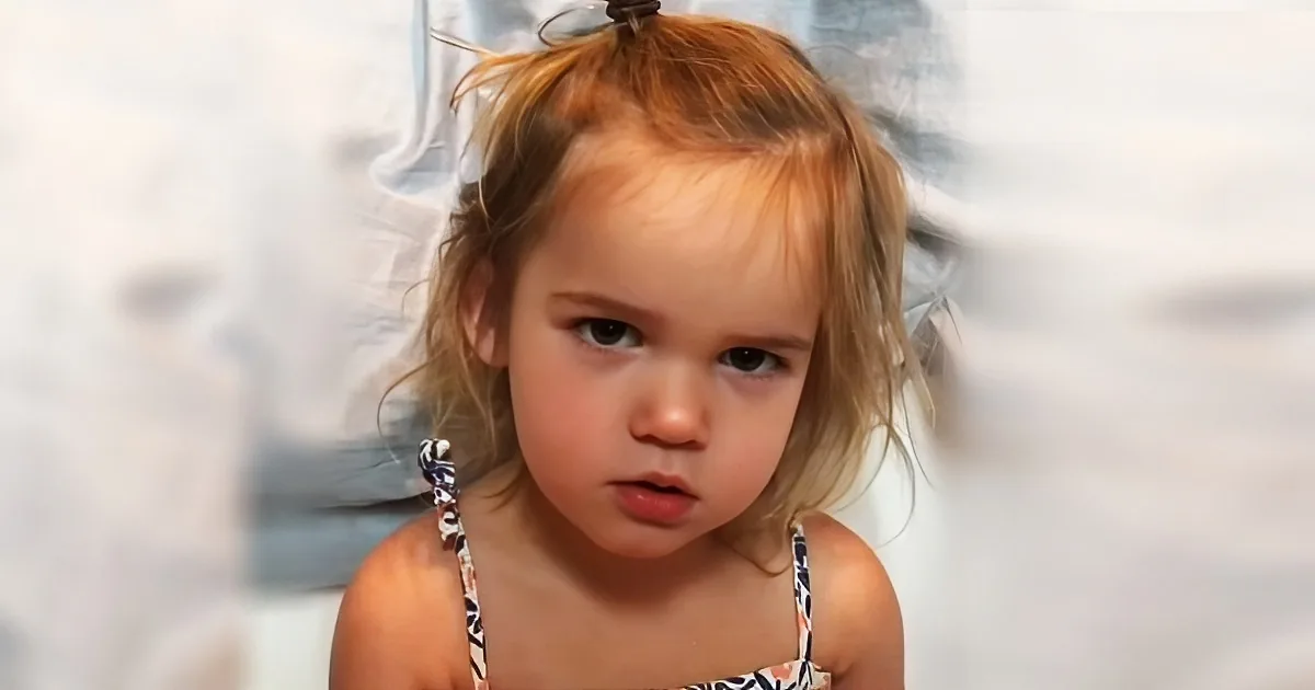 A young child with light brown hair in a ponytail, wearing a patterned top, looks off to the side with a curious expression.
