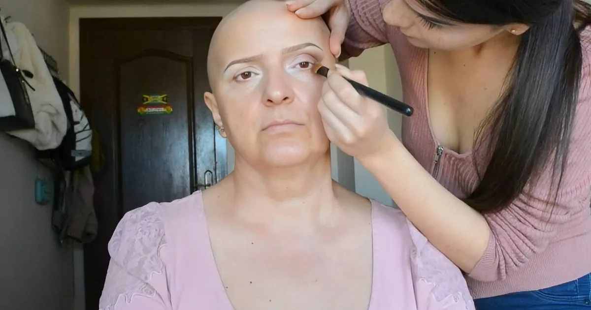 A woman applies makeup to another woman's face, both wearing pink tops.