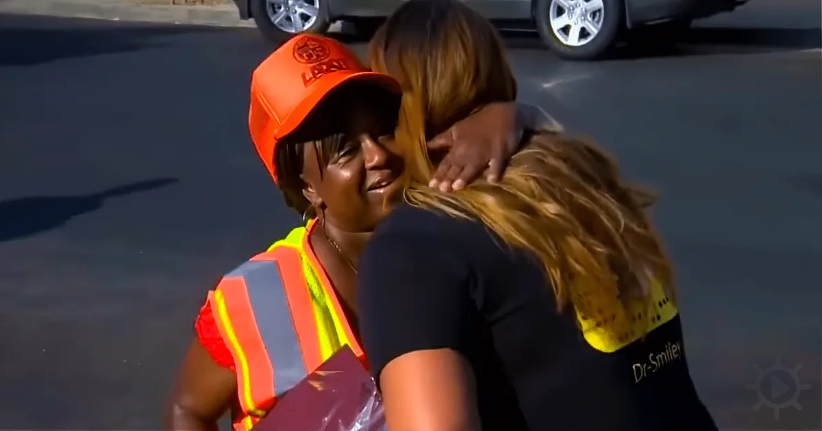 A woman in an orange hard hat and safety vest hugs another woman in a black shirt.