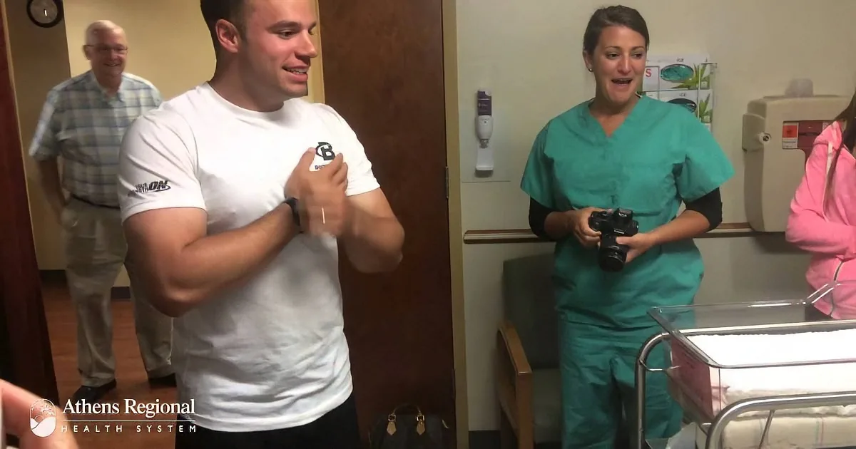 A man in a white shirt adjusts his shirt in a hospital room, with a nurse holding a camera nearby.