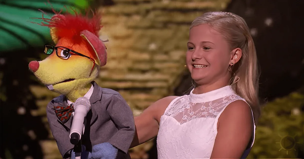 A puppet with glasses and a suit holds a microphone next to a smiling girl in a white dress.