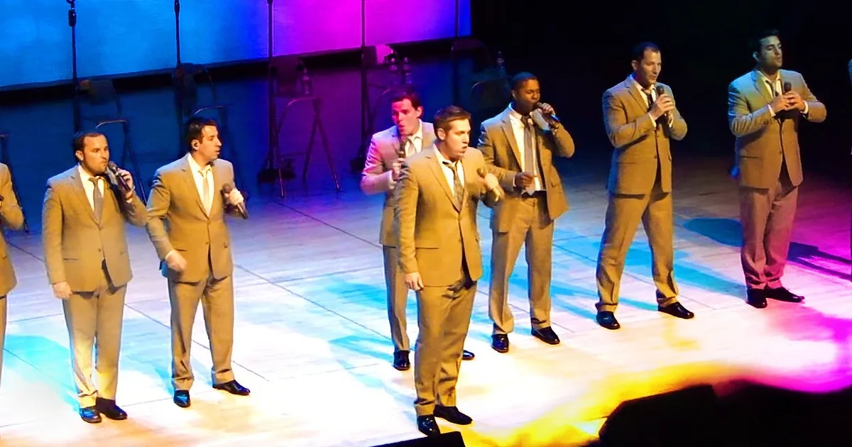 A group of men in beige suits sing on stage under colorful lights.