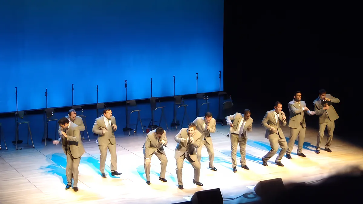 A group of men in suits performs a synchronized dance routine on a stage with blue lighting.