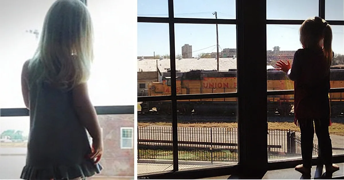 A young girl stands by a large window, gazing out at a Union Pacific train passing by.