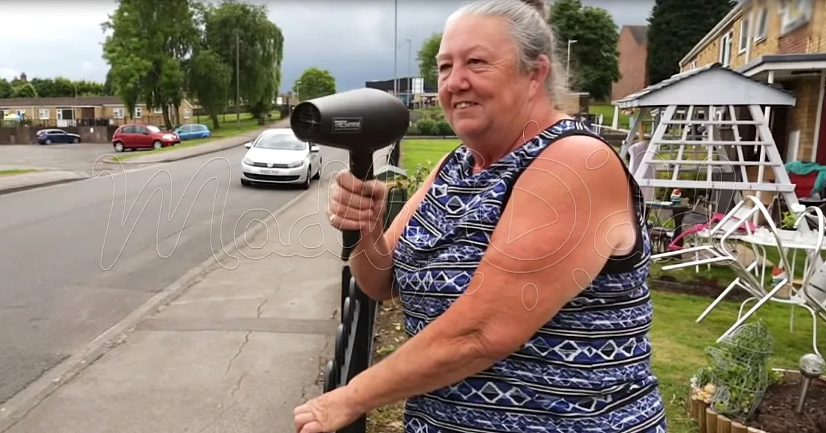 A woman holds a hairdryer, smiling, with a suburban street and parked cars in the background.