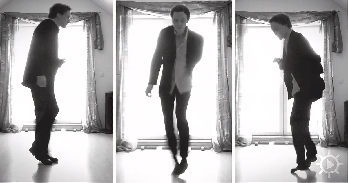 A man in a suit dances in front of a bright window with curtains.