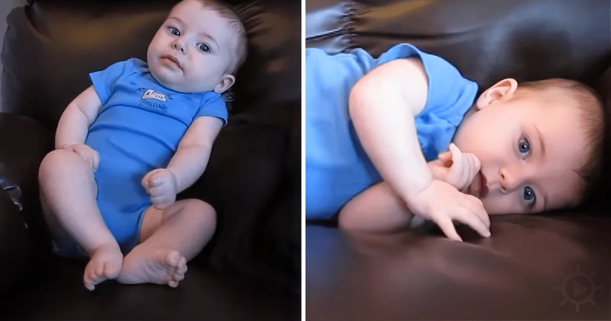A baby in a blue outfit lies on a dark surface, looking up with one hand near its mouth.