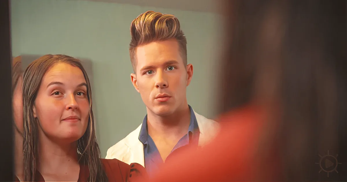 A man with a styled hairstyle looks at a woman smiling in a mirror.