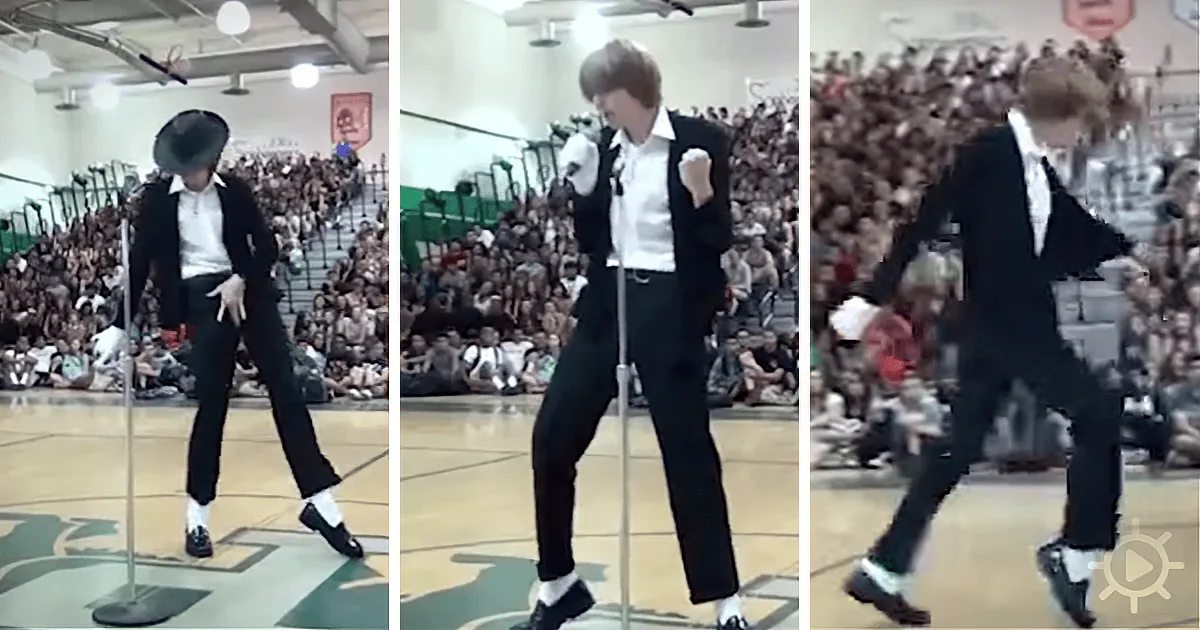 A person in a black suit and hat dances on a basketball court in front of a large crowd.