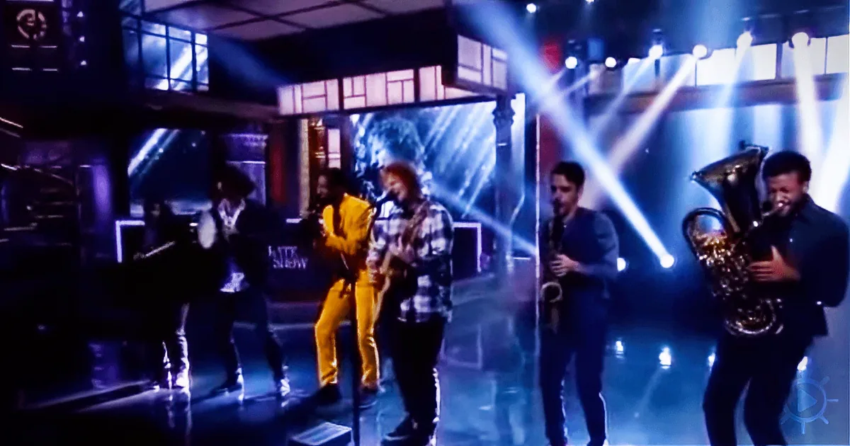 A musician in a yellow suit is surrounded by a band playing instruments on a stage with bright lights.