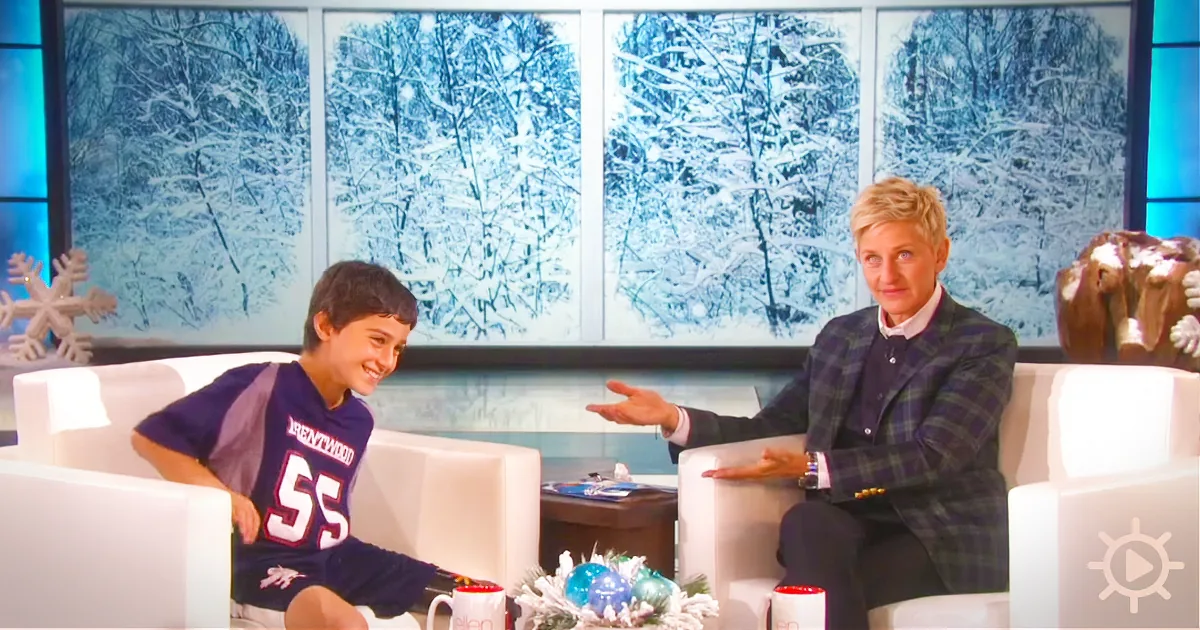 A young boy in a sports jersey sits on a white couch, smiling at Ellen DeGeneres, who gestures with her hands.