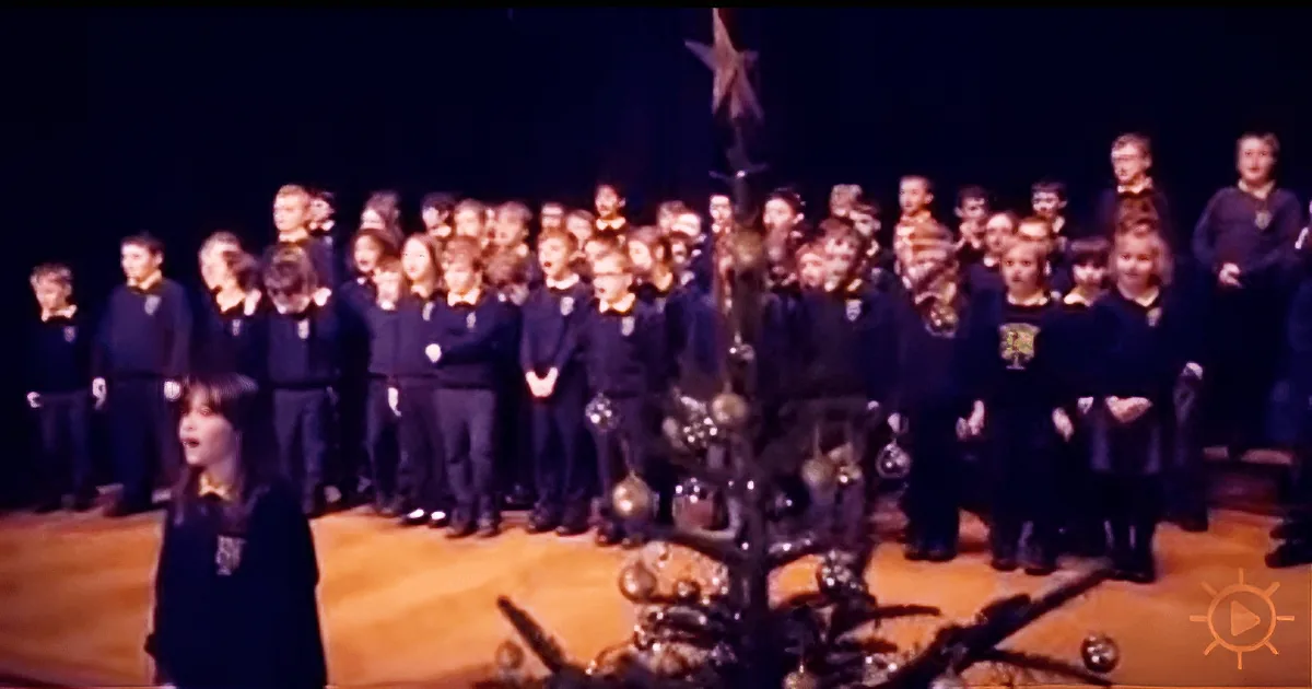 A young girl sings in front of a large choir, with a Christmas tree in the foreground.
