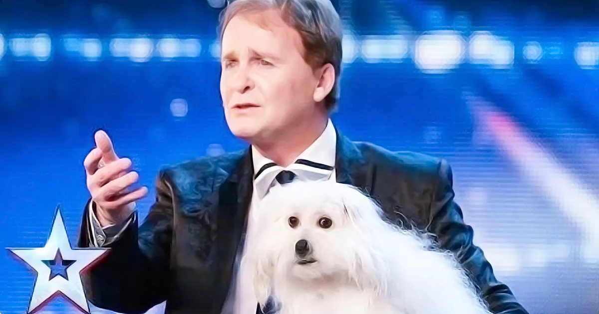 A man in a tuxedo holds a fluffy white dog, gesturing with his hand, against a blue background with a star.