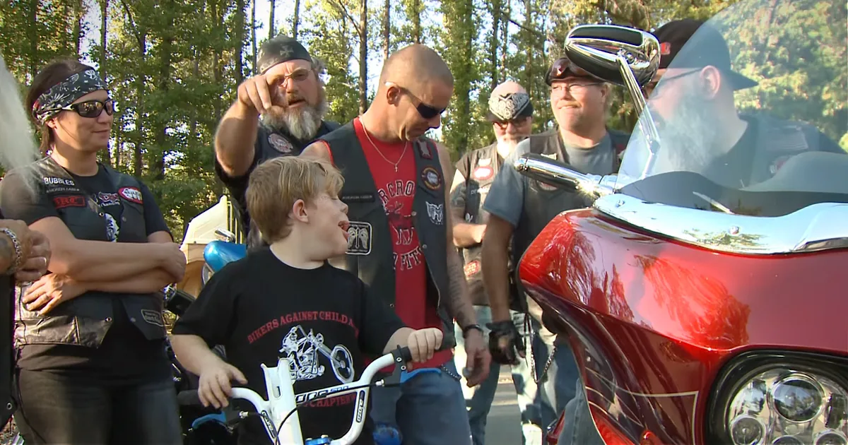 A young boy in a "Bikers Against Child Abuse" shirt interacts with a group of motorcyclists near a red motorcycle.