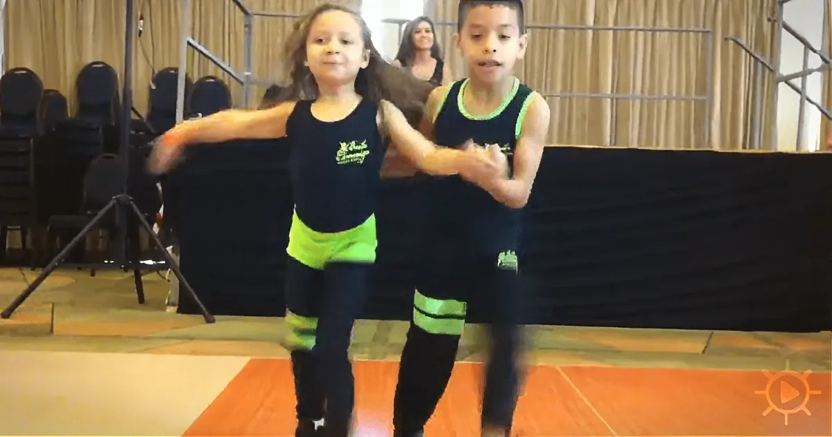 Two children in matching outfits dance on a stage, with a woman and chairs in the background.