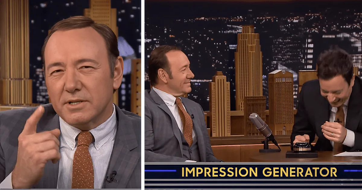 Kevin Spacey in a suit and tie, gesturing while on a talk show set with a cityscape backdrop.