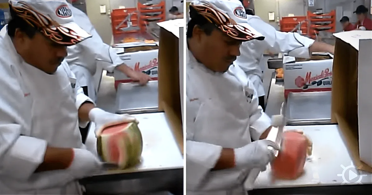 A chef in a white uniform slices a watermelon on a cutting board in a kitchen.