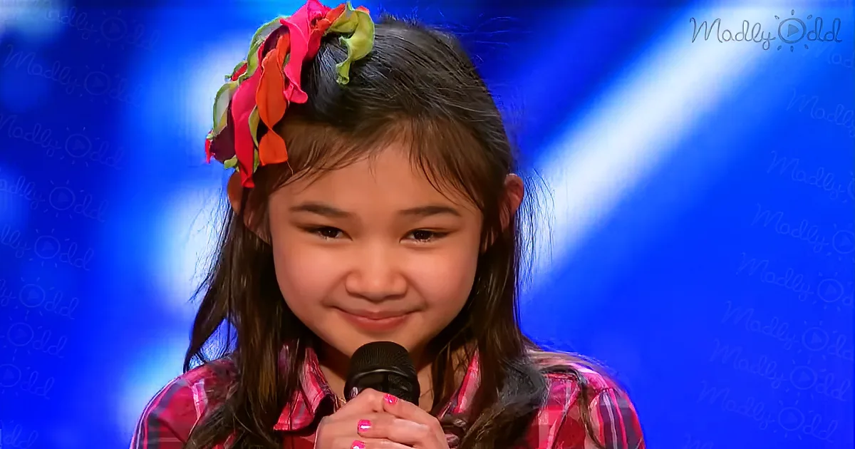 A young girl with a colorful floral headband smiles while holding a microphone on stage.