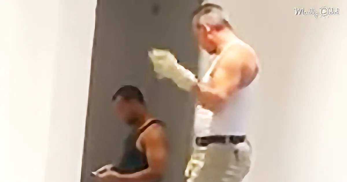 A man in a white tank top and gloves is painting a wall, with another person in the background.