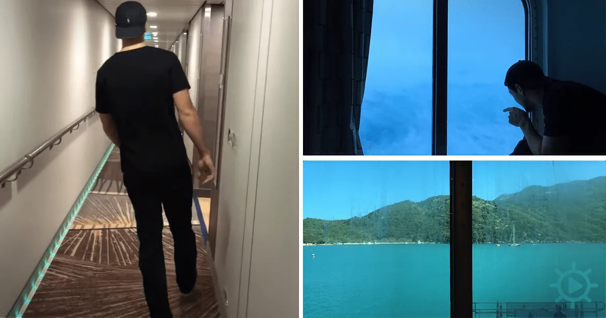 A man in a black shirt and cap walks along a ship's corridor, looking out at a scenic view of a turquoise lake and mountains.