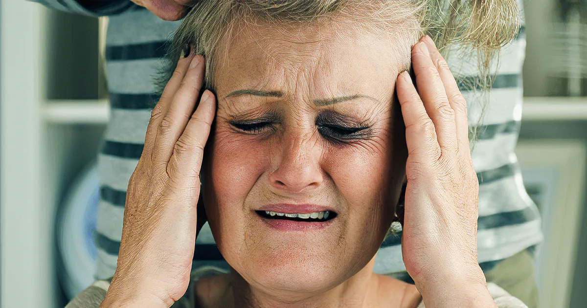 An elderly woman with hands on her head, appearing distressed, with someone behind her in a striped shirt.