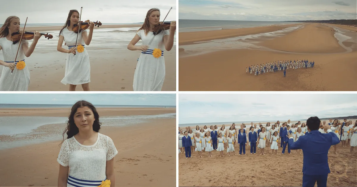 A group of people in white and blue attire stands on a sandy beach, some playing violins, others in formation, under a...