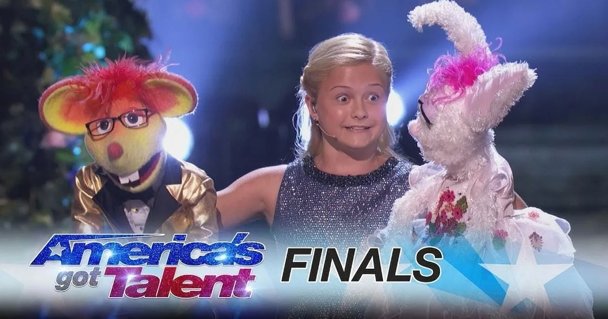 A young girl in a sparkly dress stands with two puppet characters on stage at America's Got Talent Finals.