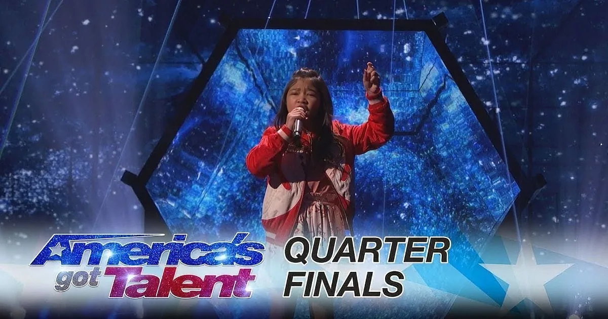A young girl in a red jacket sings passionately on stage at America's Got Talent Quarter Finals.