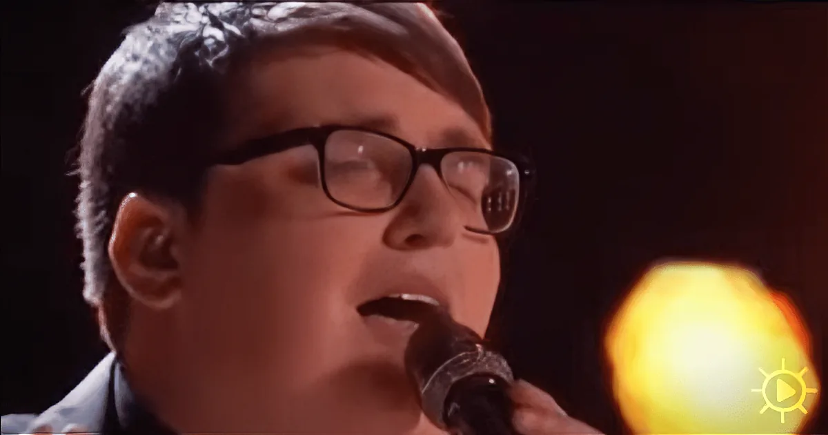 A person wearing glasses sings into a microphone, with a bright light in the background.