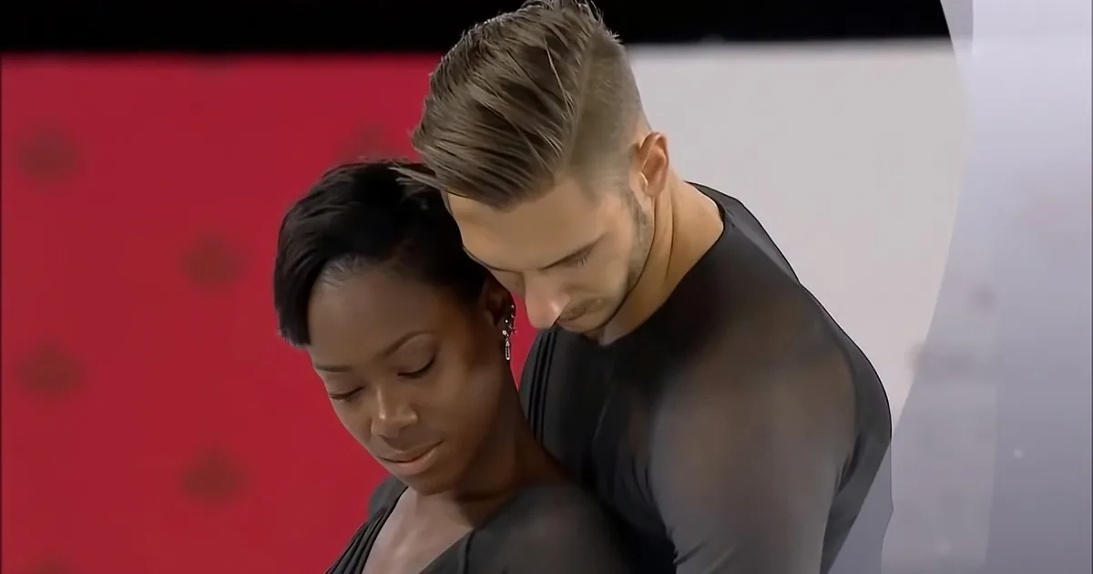 A man and woman in black outfits are closely positioned, with the woman looking down and the man leaning in.