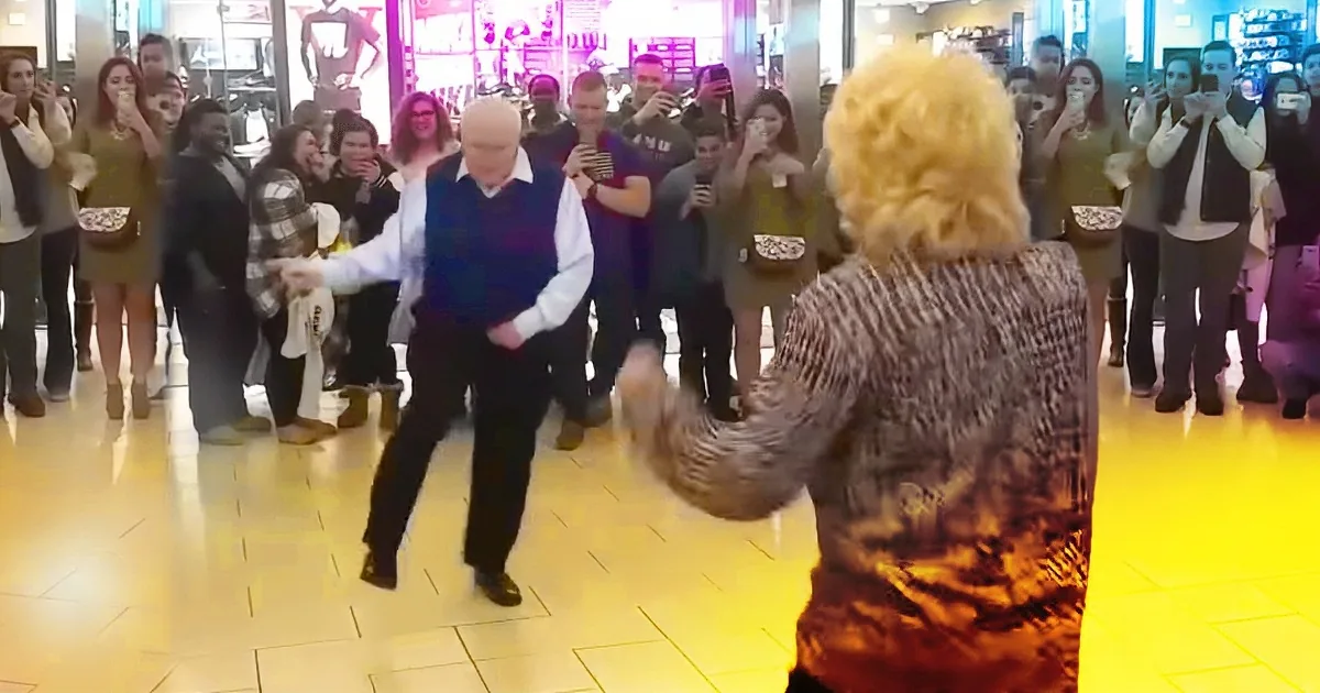 A man in a vest dances in front of a crowd of onlookers in a brightly lit indoor setting.