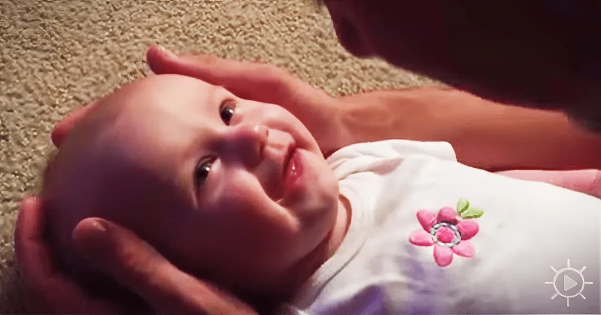 A baby in a white shirt with a pink flower is being held and looked at by a person.