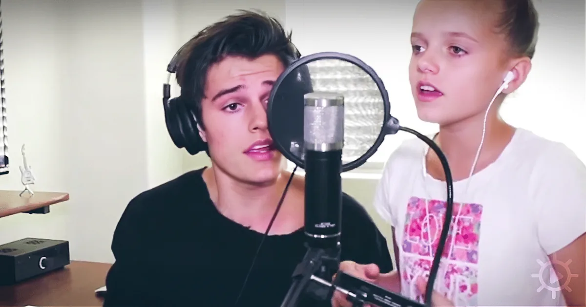 A young girl wearing earphones sits in front of a microphone, while a person with headphones listens in the background.